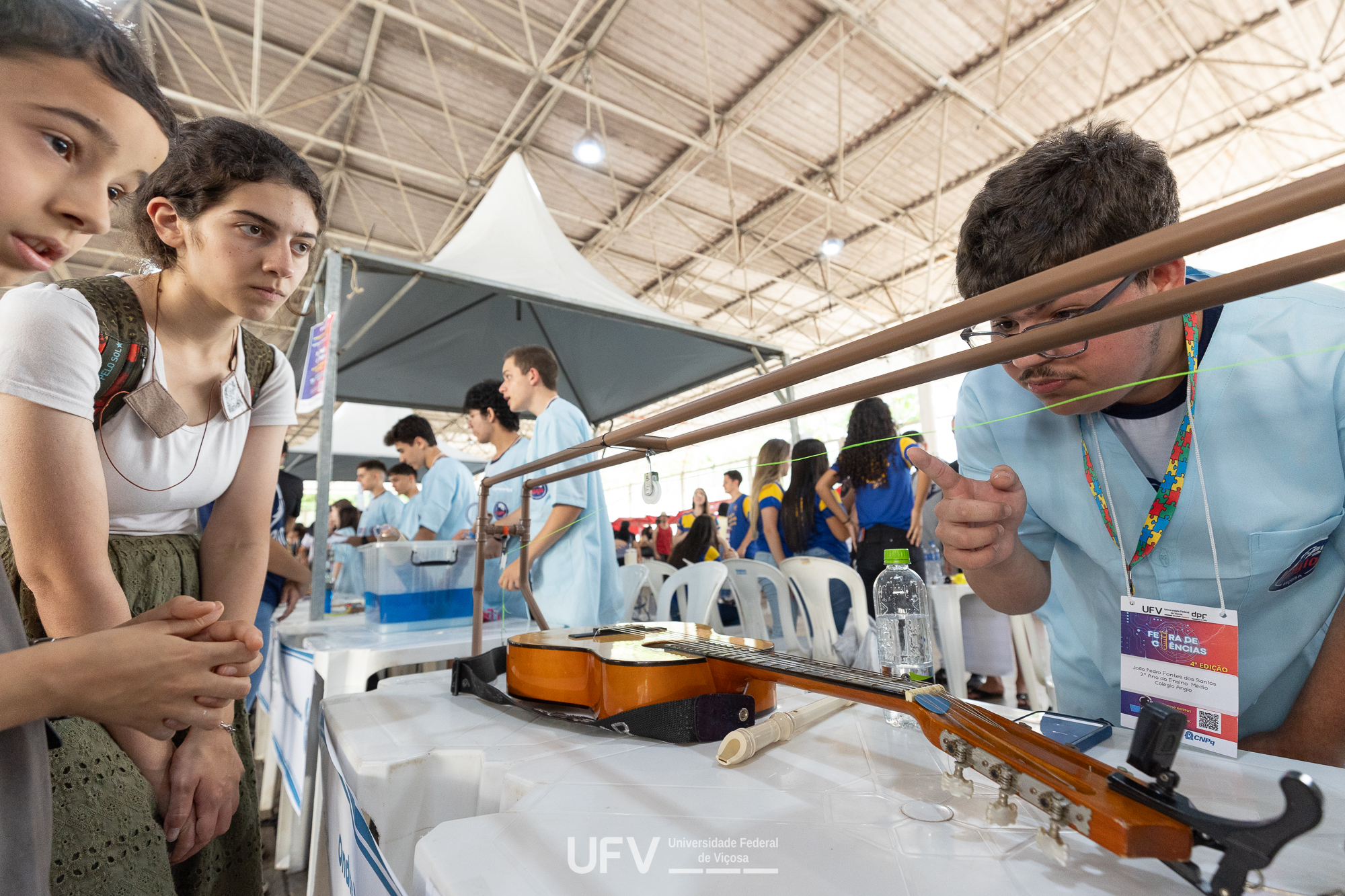 Ao centro, um violão repousa sobre uma mesa branca, com uma estrutura feita com canos de PVC montada sobre ele. À direita da mesa, um rapaz realiza um experimento com essa estrutura; à esquerda, uma moça e um menino acompanham a explicação. 