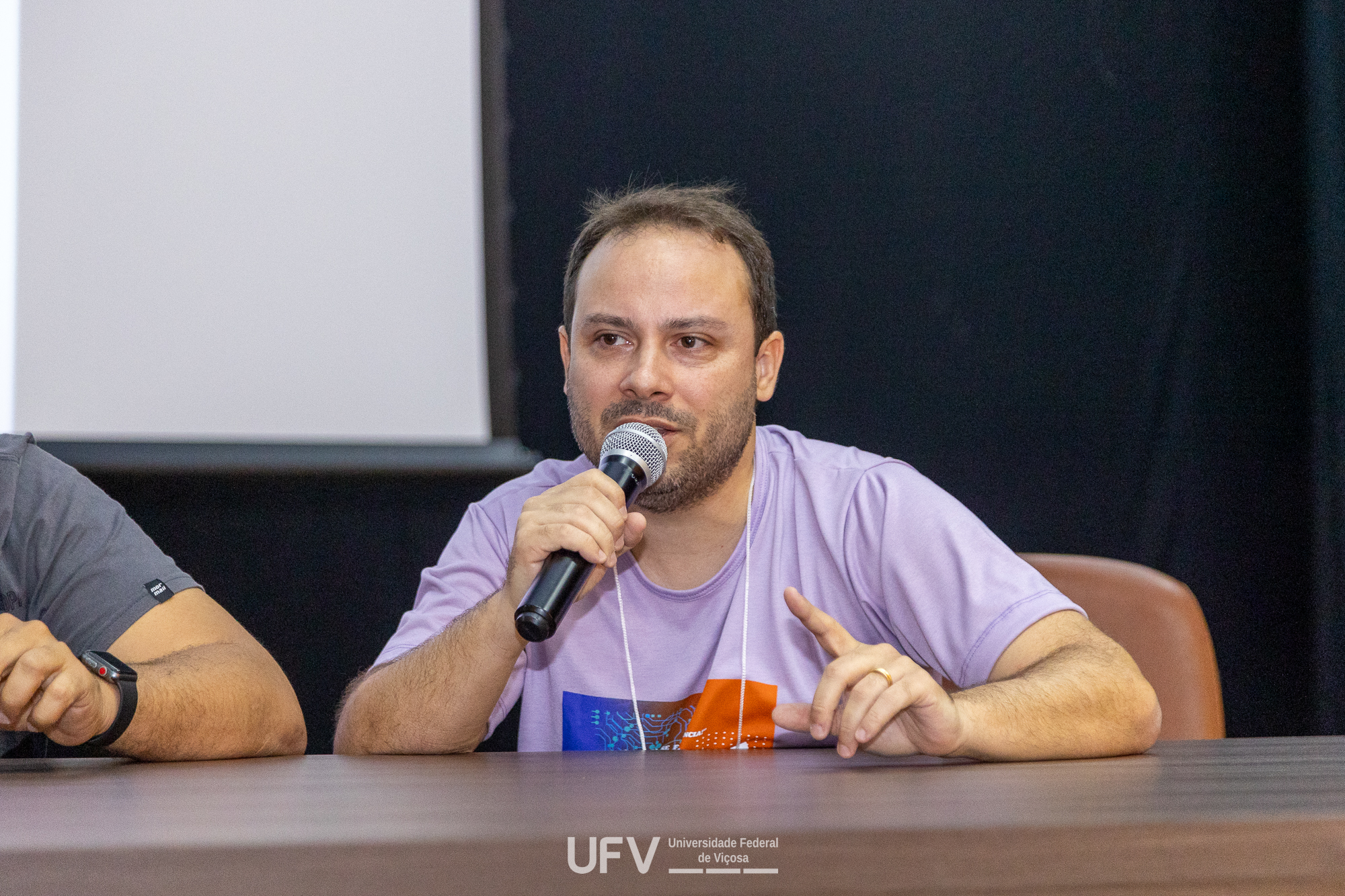 Homem branco, ligeiramente calvo, barba e bigodes por fazer e camisa roxo claro fala ao microfone, sentado a uma mesa marrom. O dedo indicador de sua mão esquerda está apontando para cima. 