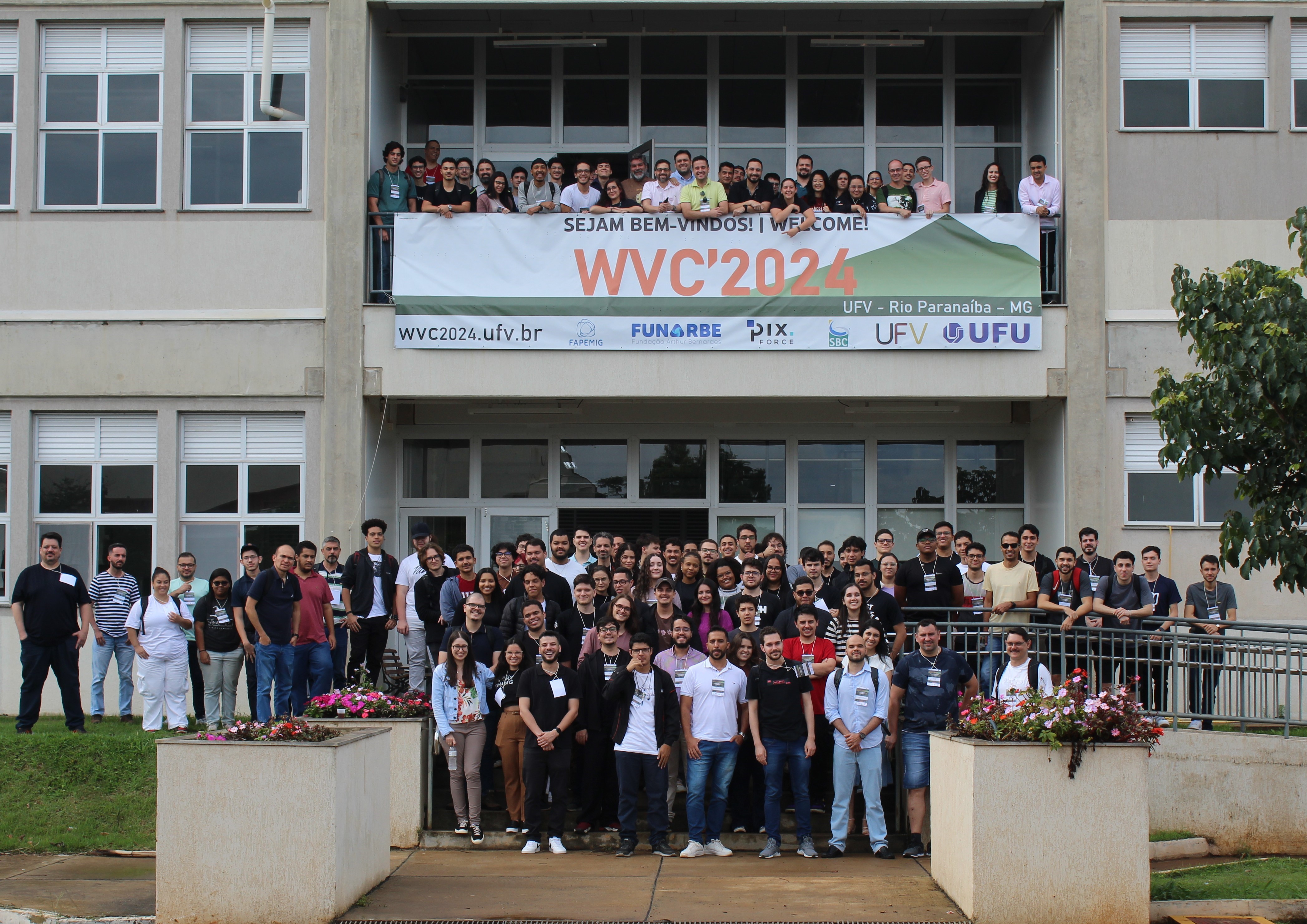 Fachada de um prédio verde claro de 2 andares. Centenas de pessoas estão de pé em frente à entrada e também em uma varanda no segundo andar. Na mesma varanda, há uma faixa onde se lê: Sejam bem-vindos! Welcome! WVC 2024. 