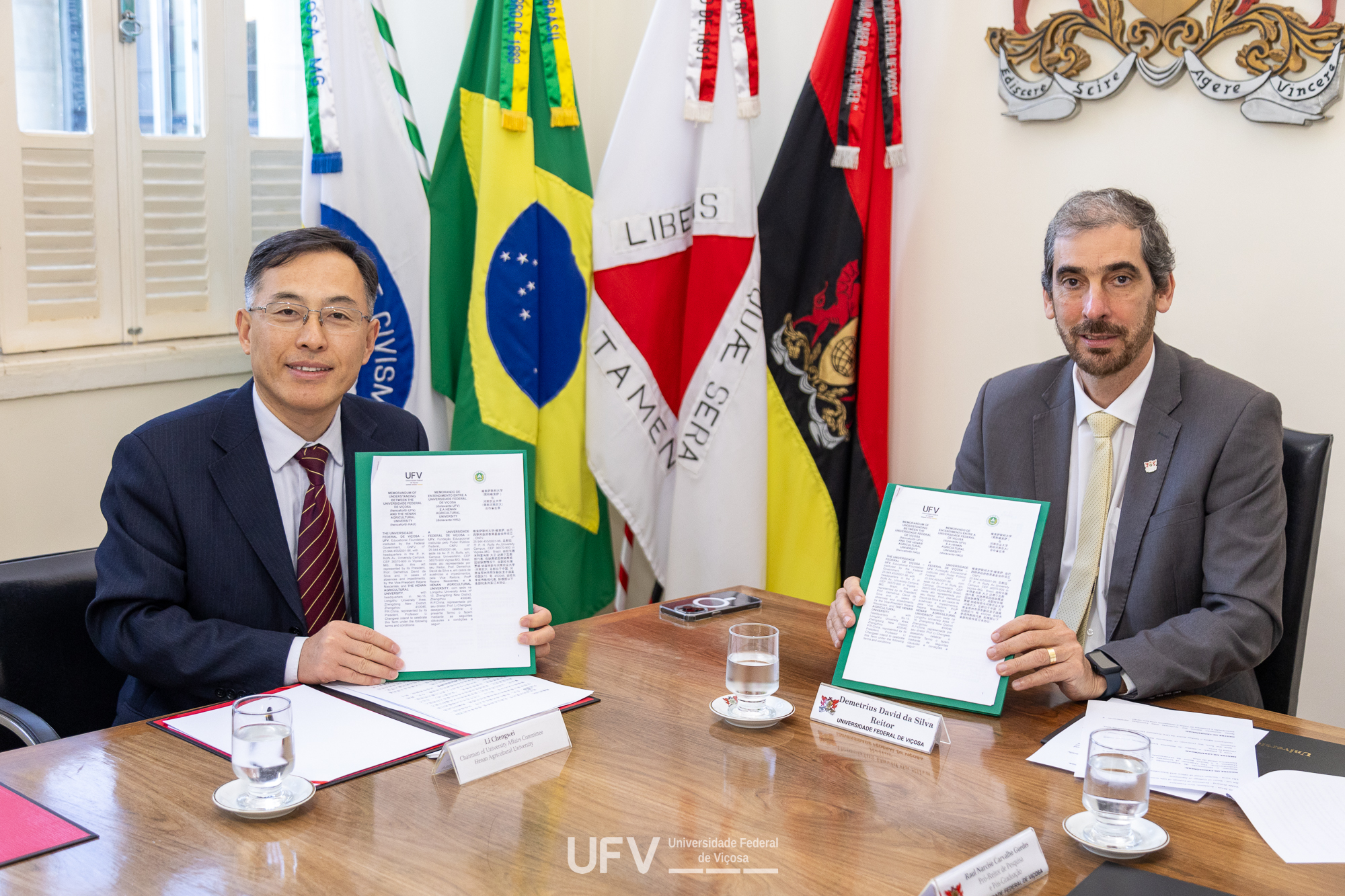 Demetrius (à direita) e Li Chegwei sentados à mesa posam segurando papel com a logo da UFV. 