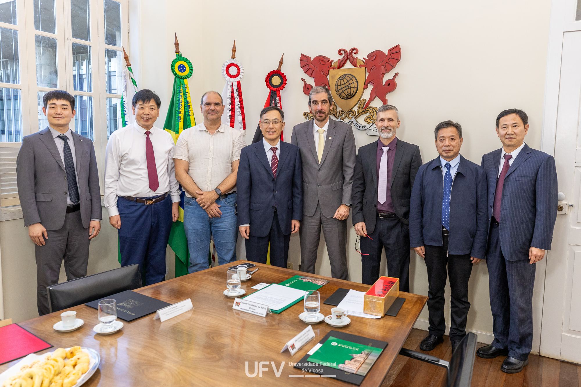 8 homens de pé lado a lado em frente a uma parede com com brasão da UFV posam para a foto. 