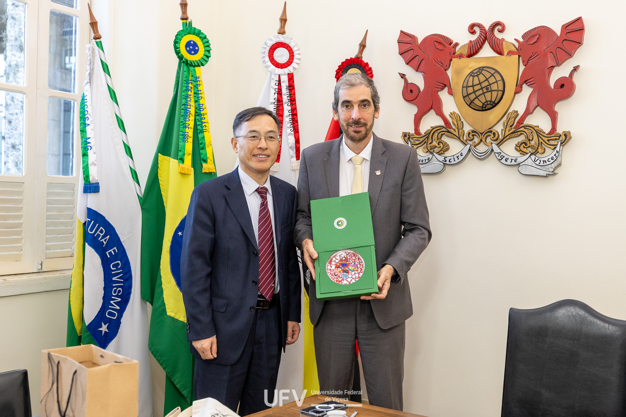 Demetrius (à direita) e Li Cegwei posam de pé para a foto. Demetrius exibe uma caixa verde com arte chinesa em vermelho.  