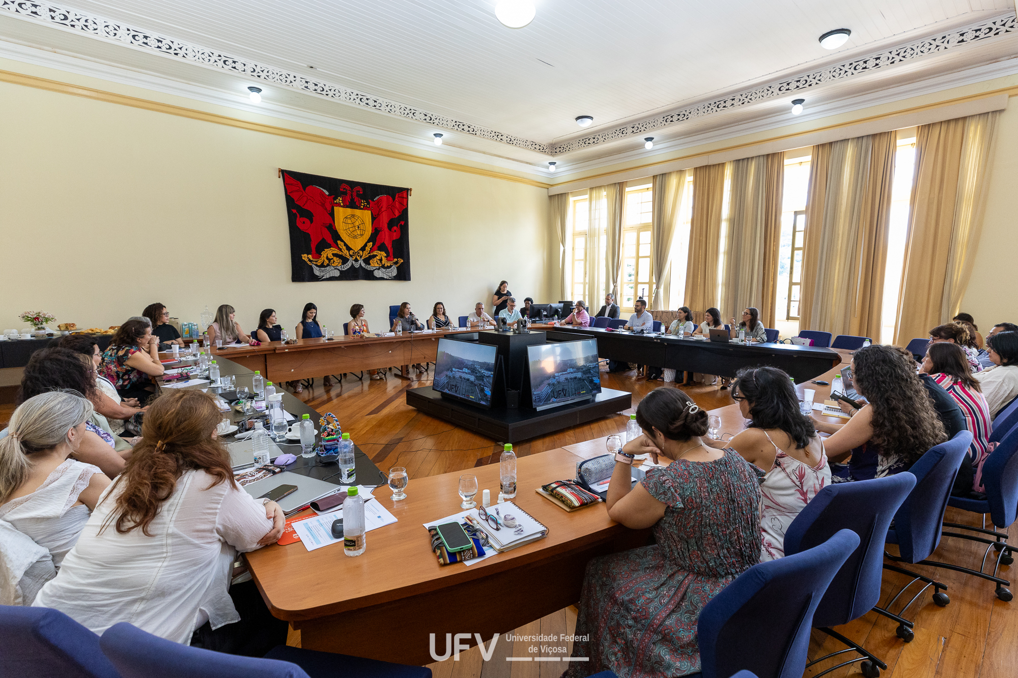 Grande sala de reuniões com piso de madeira, mesa anelar com muitas pessoas ao redor e brasão da UFV na parede ao fundo à esquerda. Ao fundo à direita, janelas grandes e altas com cortinas claras e franzidas que vão do teto ao chão. 