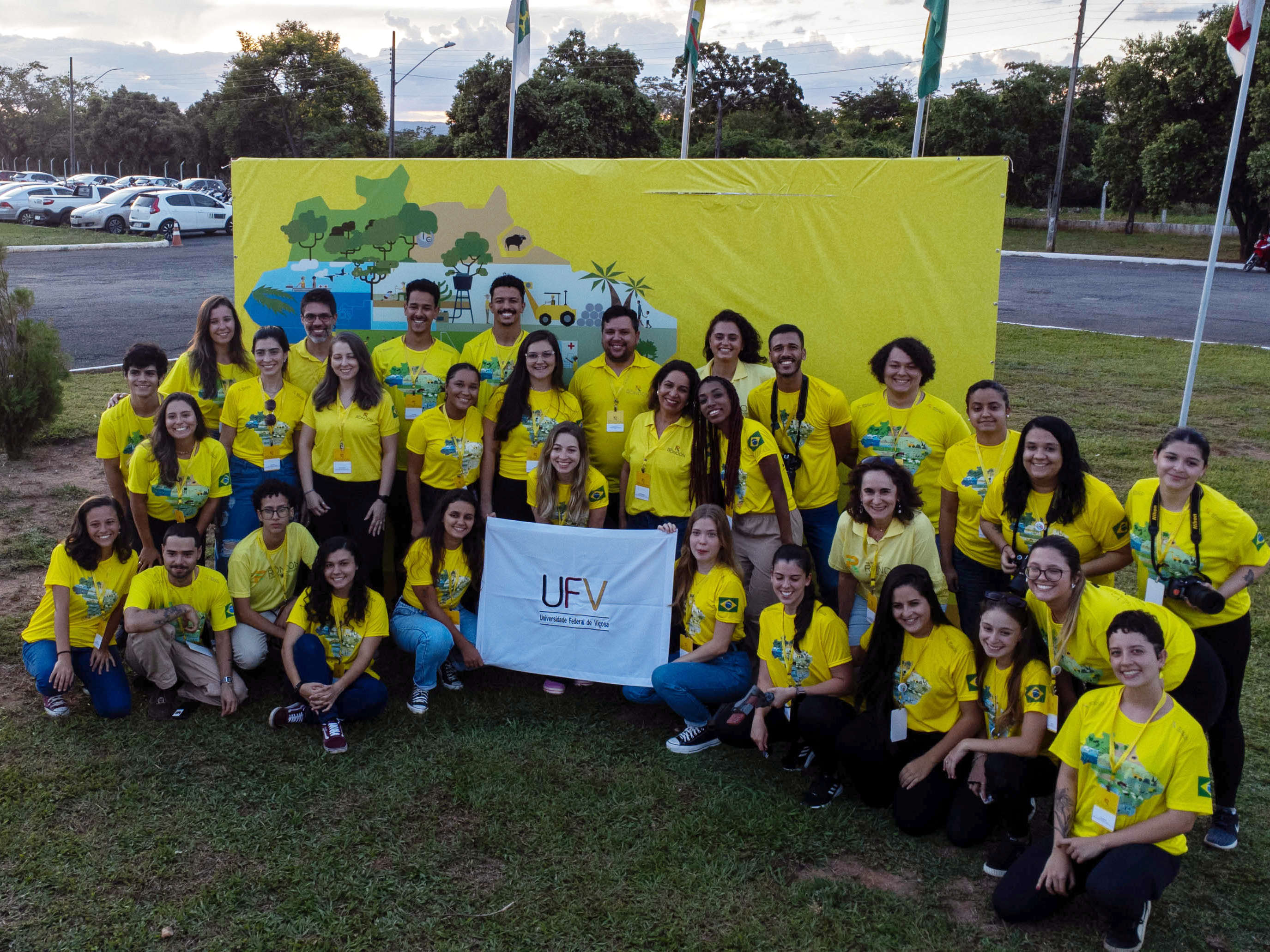 Dezenas de jovens, todos de camisas amarela com mapa do Brasil estilizado e crachás, seguram pano banco com a logo da UFV. 