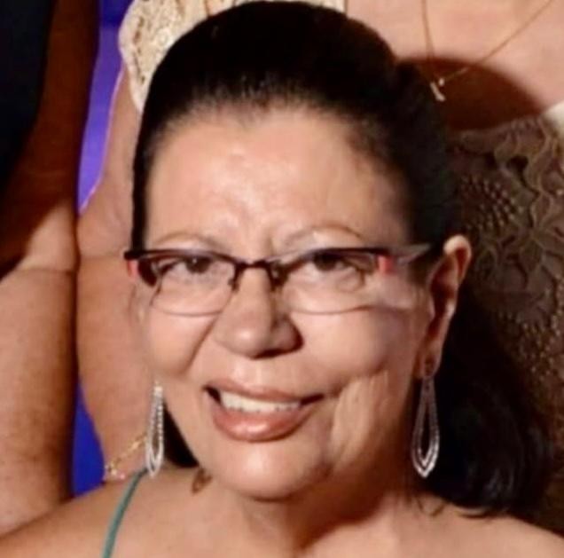 Mulher idosa de pele clara, cabelos castanhos presos, sobrancelhas finas, óculos de grau, nariz e lábios largos. 