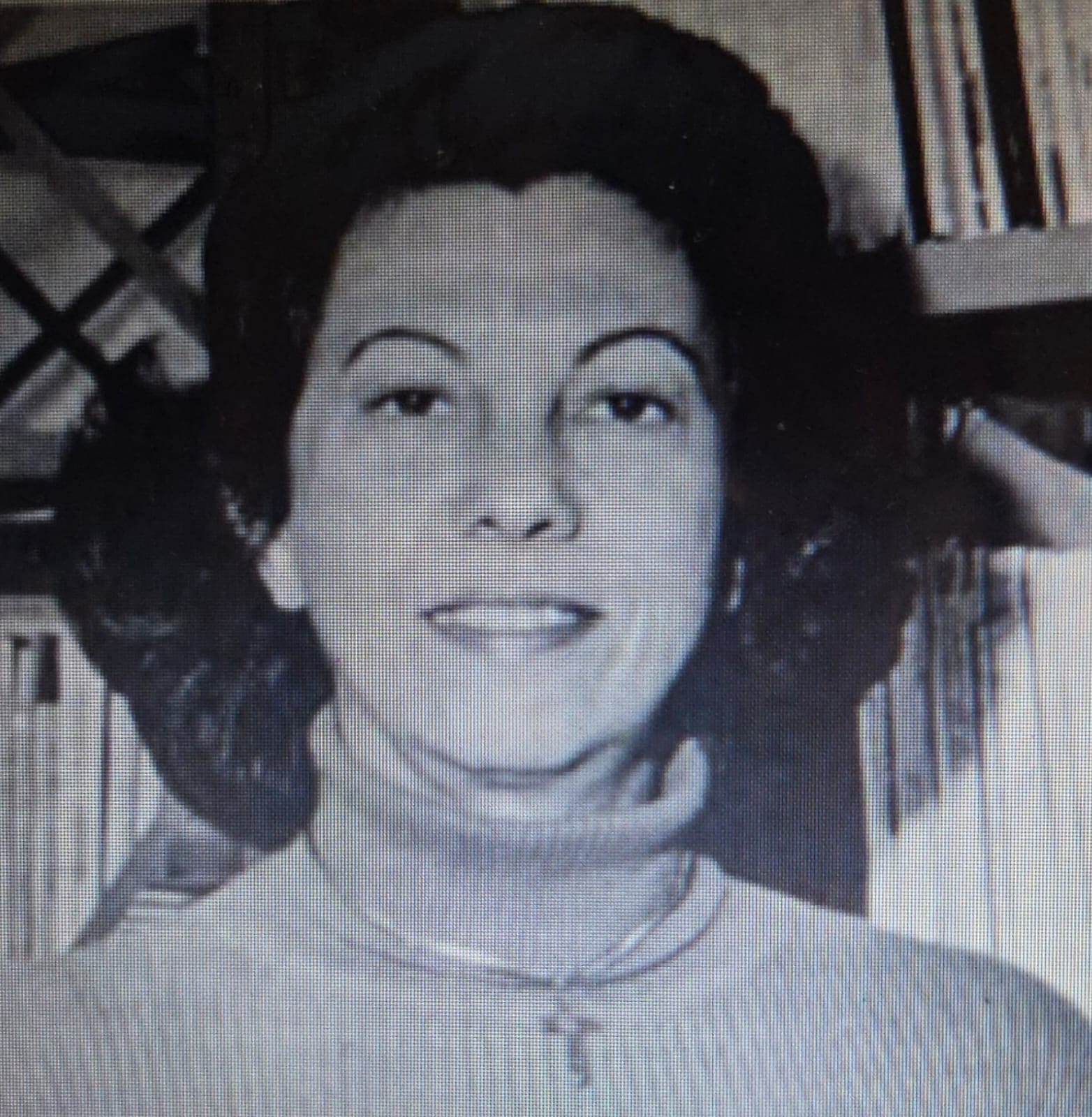 Mulher jovem magra, pele clara, cabelos pretos na altura dos ombros, sobrancelhas finas e arqueadas, crucifixo no pescoço. 