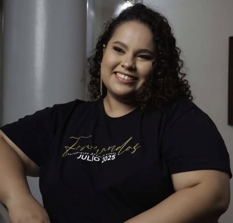 Mulher jovem, parda, cabelos pretos cacheados, olhos pretos e sorriso largo. Ela usa camisa dos formandos de julho 2025. 