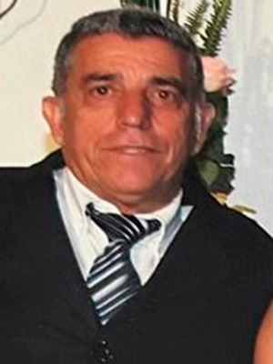 Senhor idoso, pardo, cabelos levemente grisalhos penteados para trás, sobrancelhas e olhos pretos, terno preto. 