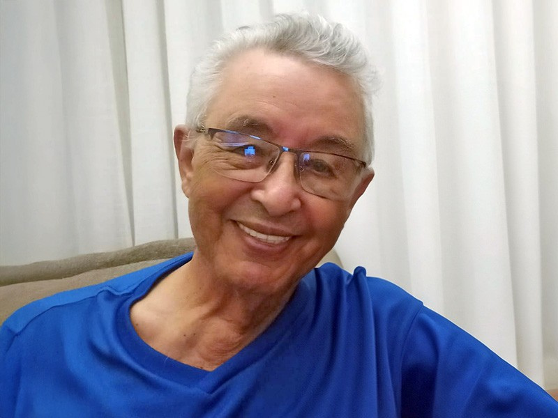 Homem claro, magro, sorridente, cabelos brancos, olhos oblíquos, nariz levemente adunco, óculos e camisa azul escuro. 