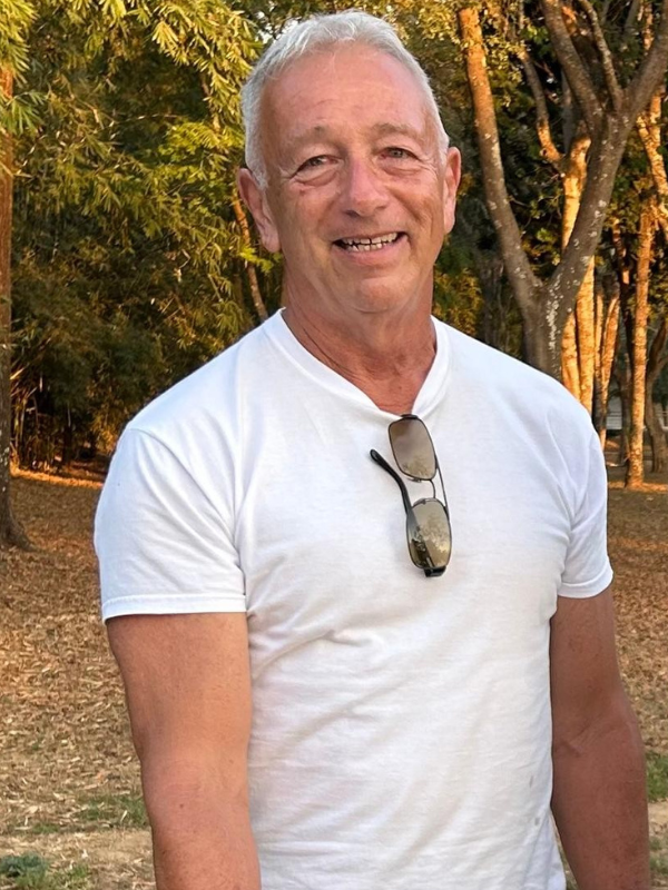 homem branco, magro, alto, cabelos grisalhos e lábios finos, em área gramada com árvores ao fundo. Ele sorri. 