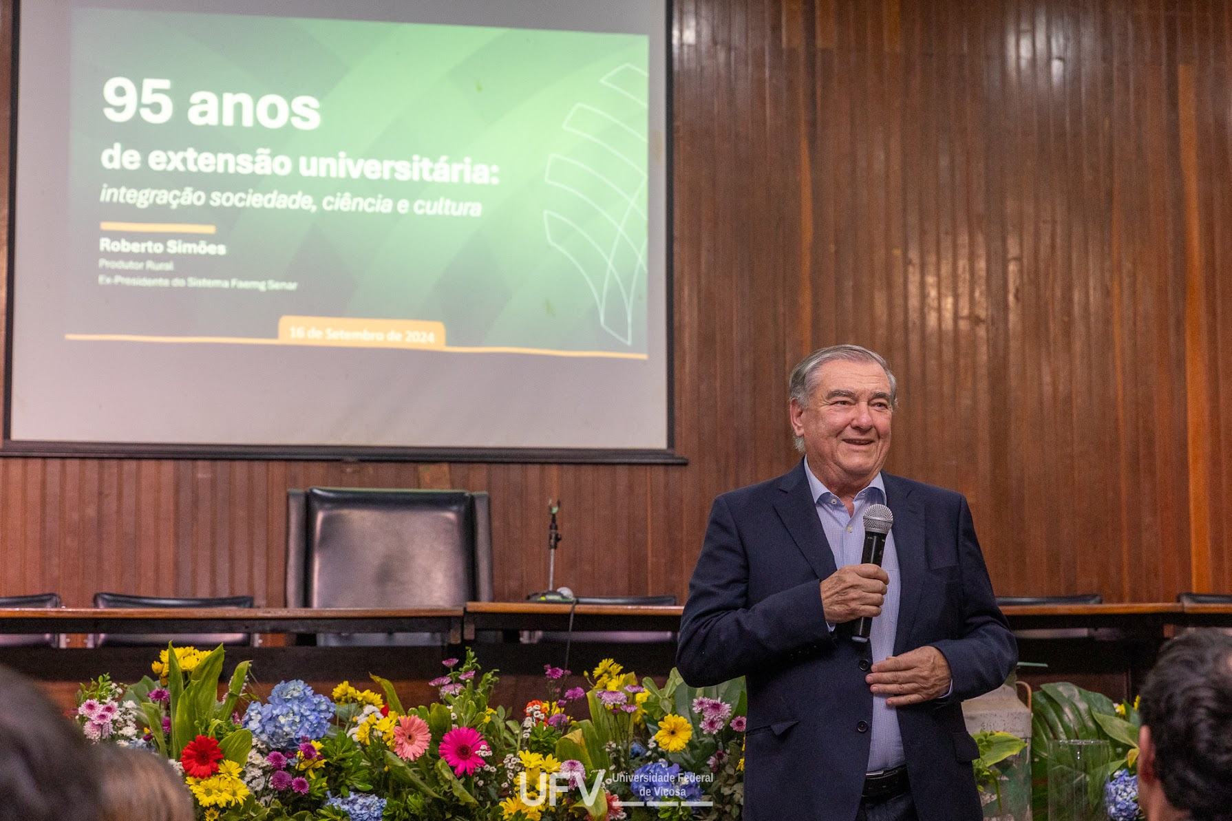 Senhor idoso branco, cabelos lisos grisalhos e sobrancelhas grossas, de pé em frente a um telão e segurando um microfone. 