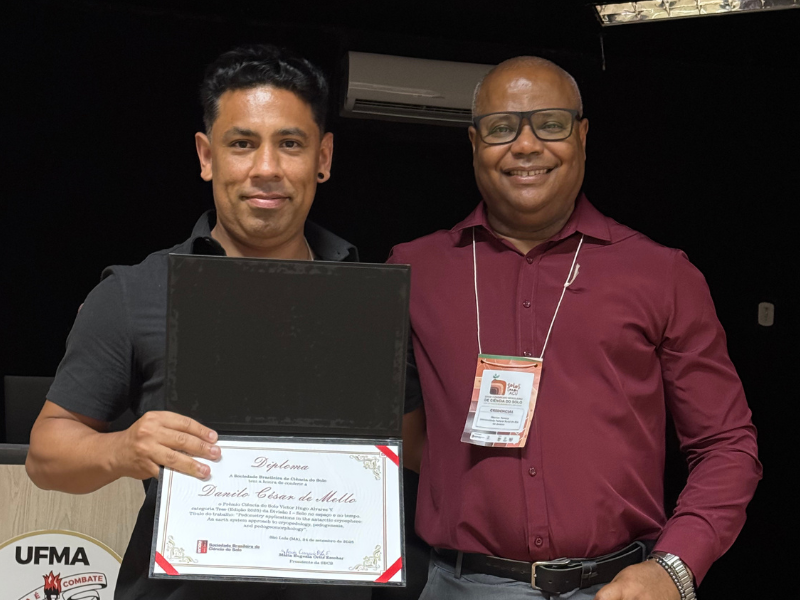 2 homens de meia-idade de pé lado a lado; o da esquerda segura um certificado em frente ao corpo. 