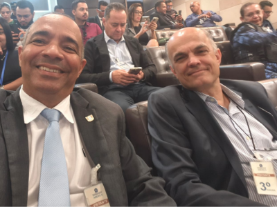 Dois homens sentados num auditório sorriem para a foto. O da esquerda usa gravata, o da direita não.  Dois homens sentados num auditório sorriem para a foto. O da esquerda usa gravata, o da direita não.
