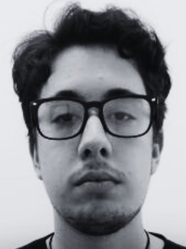 Rapaz de pele clara, cabelos pretos ondulados curtos, sobrancelhas grossas, óculos de aro preto. Ele olha para nós sério. 