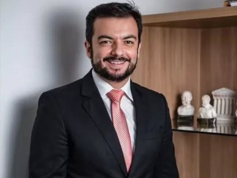 Homem jovem, branco, testa alta, cabelos barba e bigode pretos. Ele olha para nós e sorri. Usa terno preto e gravata rosa. 